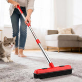 Person using a red broom on a carpet with a cat nearby in a living room setting.